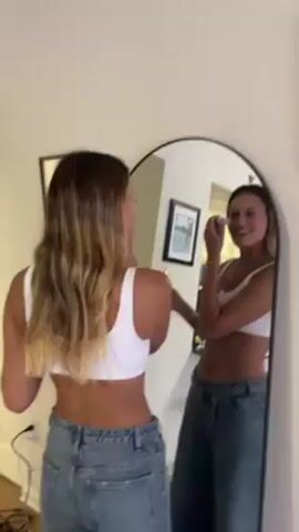 A woman with long brown hair is standing in front of a rounded mirror, applying makeup. She's wearing a white crop top and baggy blue jeans. In the reflection, she's smiling as she applies makeup to her face. Someone off-camera holds up a large silver sword, briefly obscuring the view of the mirror. The room is otherwise empty, with a white door in the background and a framed picture on the wall.