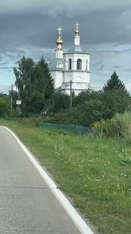 The video shows a white church with gold domes in a rural setting. The church is surrounded by trees and greenery. A road runs along the left side of the frame, with a white line marking its edge. The sky is cloudy and dark.