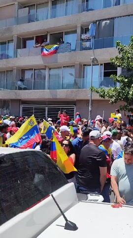 The video depicts a lively street gathering. A large group of people are crowded together, waving flags of Venezuela. Some are wearing hats and clothing with Venezuelan flag designs. A woman in a black top holds a flag high, while others around her are cheering and celebrating. The background features a tall, multi-story building with balconies, some of which display the Venezuelan flag. A few people are visible on the balconies, observing the crowd below. A couple of parked cars are in the foreground, partially obstructing the view. The scene is brightly lit, suggesting it is daytime.