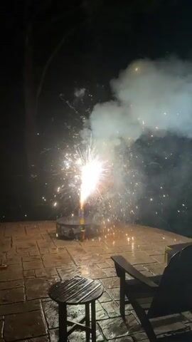 The video captures a nighttime scene of fireworks being set off on a paved patio. The fireworks are emitting sparks and smoke, illuminating the dark sky. A small round table with a striped top is visible in the foreground, along with a dark-colored chair positioned to the right. The ground appears wet, suggesting recent rain. The fireworks are the central focus, creating a bright, explosive display.