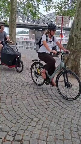 The video begins with a close-up of a red cargo bike with a black cover being ridden by a person wearing glasses and a grey t-shirt. The bike is on a cobblestone path. The camera then pans to the right, showing another person on a bicycle with a backpack. In the background, there is a bridge, trees, and buildings. As the video continues, more people are seen walking on the path. The cargo bike and the other bicycle move further down the path, with the cargo bike in front. The camera then pans to the right again, following two people walking away from the camera. The video ends with a view of the path lined with trees and several people in the distance.