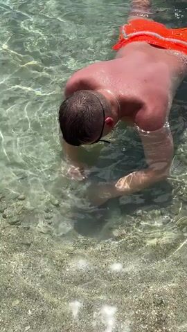 A man wearing bright orange swimming trunks and dark goggles is floating on the surface of clear water, looking down with his face submerged. He uses his hands to explore the rocky, sandy bottom of the shallow water. The sun creates shimmering patterns on the surface of the water and illuminates the seabed.