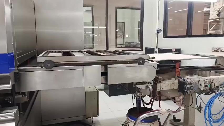 A conveyor belt moves a large rectangular object with multiple compartments across a factory floor. The object appears to be a frame or a tray. The factory environment is industrial, with stainless steel machinery and bright fluorescent lighting. The camera is static, capturing the object in motion.