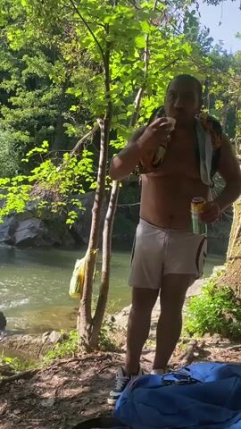 A man with a towel over his shoulder stands near a river, holding a can and an apple. He is wearing white shorts and sneakers. In the background, the river flows over rocks surrounded by trees. The second part of the video shows a close-up of another person's face, and then a blurry shot of legs.