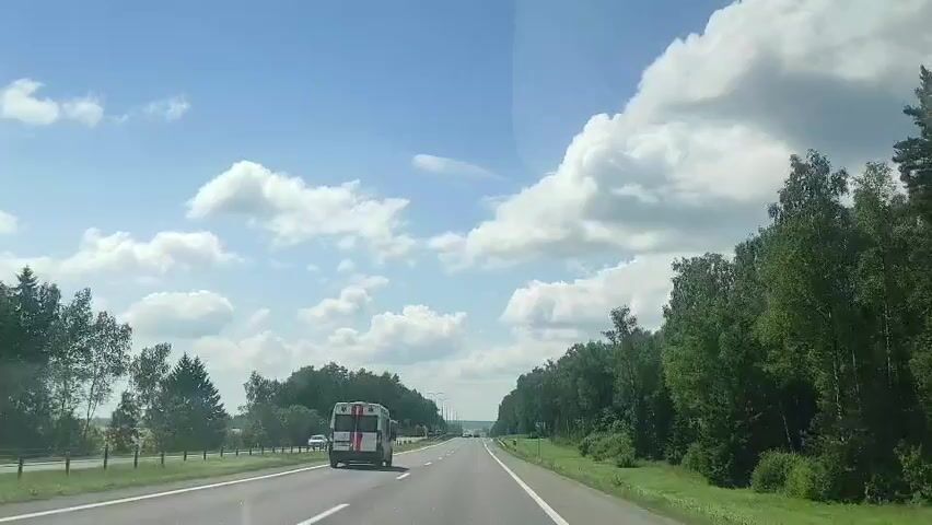 The video shows a view from the passenger seat of a vehicle traveling on a highway. The sky is blue with fluffy white clouds. On the right side of the highway, there is a dense forest. On the left side of the highway, there are fields and trees. In the distance, a white van with red markings is visible, traveling in the same direction. Other vehicles are also present on the highway. The road has lane markings and appears to be in good condition. The overall scene suggests a bright, sunny day.