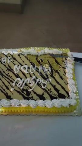 The video displays a close-up of a rectangular cake with yellow frosting, decorated with black stripes and white lettering. The cake appears to be part of a celebration, possibly a birthday, as indicated by the number "18" written on it in white icing. Next to the cake, a slice has been cut and is partially visible. Further down the table, there are two long, cylindrical cakes, also decorated with frosting and fruit. Cutlery, including forks and knives, is laid out on the table, along with stacks of plates and lit candles in glass containers. The table is covered with a white tablecloth. The overall scene suggests a festive gathering.