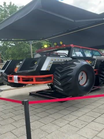 The video shows a monstrous truck with large tires and an orange frame. The truck is covered by a black tent. The truck has flames on its side and a silver front end with gold rivets. There are also yellow lights on top of the truck. The truck is on display outdoors on a paved area with a red rope barrier in front of it.