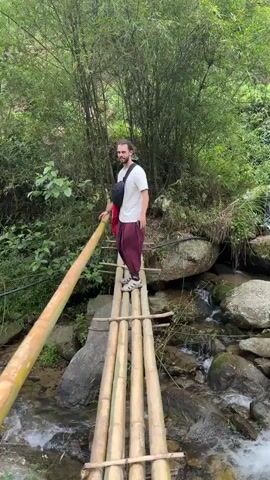 A man with long dark hair tied in a bun, wearing a white t-shirt, maroon pants, and sandals, walks across a narrow bamboo bridge over a rocky stream. He has a black messenger bag and a red bag slung over his shoulder. The bridge is constructed of multiple bamboo poles lashed together. Lush green vegetation, including bamboo trees, surrounds the stream. The stream flows over and around large rocks. The man is looking around as he carefully walks across the bridge.