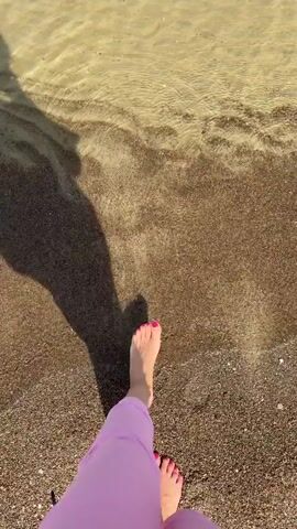 The video shows a person walking into the water. The person is wearing pink pants. The person’s foot with red toenail polish is visible as it enters the water. The water is shallow and the bottom is sandy. The water is clear enough to see the sandy bottom. The shadow of the person holding the camera is visible. The sky is blue with a few clouds on the horizon.