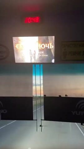 The video shows the interior of a vehicle, likely a bus, looking forward through the windshield. The time displayed on a digital clock at the top is 20:48. A screen mounted above the windshield displays the title "СЕЛАЯ НОЧЬ" (Selyaya Noch, meaning "Gray Night" or "Pale Night") in large, stylized text, with smaller text below that appears to be "Techno Deep House Cover." To the right of this screen is a yellow sign with a pictogram of a person wearing a seatbelt and text that reads "ПРИСТЕГНИТЕ РЕМНИ!" (Pristegnite remni!, meaning "Fasten your seatbelts!").

The windshield offers a view of a road stretching into the distance under a twilight sky. The sun is setting, casting a warm orange and yellow glow across the horizon. There are a few cars visible on the road ahead. The landscape outside appears to be relatively flat with some vegetation.

The overall impression is of a journey in progress during the evening. The presence of the seatbelt sign and the nature of the video suggest that this is a passenger vehicle. The music title on the screen hints at the audio content accompanying the visual. The time of day and the lighting indicate it is dusk or early night.