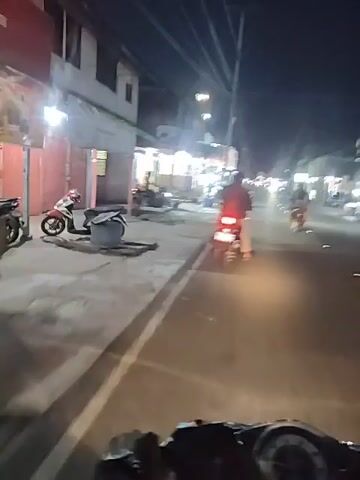 A nighttime video shows a street scene with several motorcycles parked along the side of the road and others being ridden. Buildings with lights are visible on the left side of the street, suggesting a commercial or residential area. The camera appears to be mounted on a moving motorcycle, as indicated by the motion blur. One motorcycle is clearly visible in the foreground on the right side, with its taillight illuminated. Another motorcycle is seen in the middle ground on the left, with a rider. The overall atmosphere is that of a busy street at night, likely in Southeast Asia given the prevalence of motorcycles.