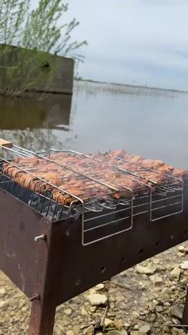 The video shows meat being grilled over charcoal on a rusty barbecue grill. The grill is positioned by a body of water with a cloudy sky in the background. In the near background, there is a small, weathered structure with foliage near the water's edge. The ground around the grill is covered with small stones. Smoke is rising from the grill, indicating active cooking.