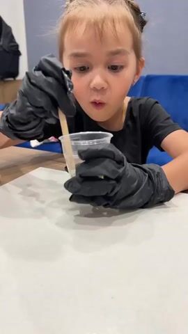 A young girl with pigtails, wearing a black t-shirt and black gloves, is sitting at a table and stirring a clear plastic cup with a wooden stick. The cup appears to contain a mixture of liquids and possibly glitter. The background shows a wooden table, a blue couch, and a portion of a black bag. The girl is looking intently at the cup as she stirs.