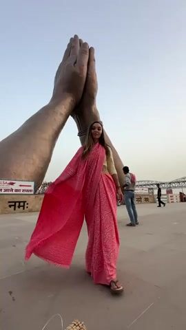 A woman in a pink saree is standing in front of a large sculpture of praying hands. She is holding out the end of her saree, which is billowing in the wind. The scene is set on a large, open, grey concrete platform. There is a sign in the background with writing in a foreign language on it, as well as other people walking around. The sky is blue.
