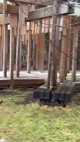 The camera pans around a wooden structure in a park. The structure has a series of wooden beams and planks, with a white canopy stretched over the top. Several piles of wooden planks rest on the grassy ground. The grass is a dull green. A patch of snow is visible in the distance. The wood of the structure appears old and weathered, and some of the beams are slightly askew. The structure appears to be a playground or art installation. There are no people visible in the video.
