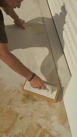 A person is kneeling on a marble floor, holding a grout float with a sponge. They are using the float to spread grout into the seams between marble tiles. The floor is light-colored with brown and green veins. A white corrugated wall is visible on the right side of the frame. The person is wearing a dark t-shirt and bracelets on their left wrist. They are focused on their task, with their head bent down. The lighting suggests an indoor environment, possibly a home or a commercial space.