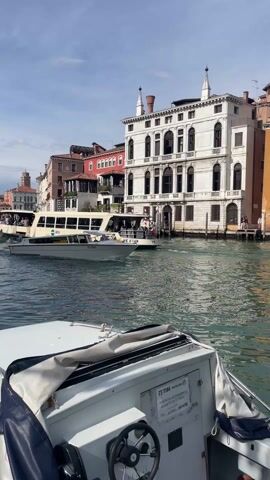 The scene captures a view of the Grand Canal in Venice, Italy, from a boat moving along the water. A white boat with a blue cover and steering wheel dominates the foreground, with a partially visible sign mentioning "TIM." Beyond the boat, a Venetian water bus, filled with passengers, glides through the canal. To its right, a smaller, sleeker white boat cruises by. The backdrop showcases a blend of Venetian architecture, featuring buildings in various colors, including white, red, and tan, lining the canal. A church tower can be seen in the distance. The water reflects the sky, which is partially cloudy.
