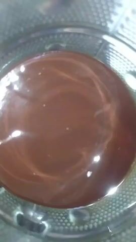 The video shows a close-up, overhead view of a dark brown liquid in a clear glass or container. The liquid appears to be smooth and slightly viscous, with reflections of light on its surface creating swirling patterns. The container has a textured rim or base visible around the liquid. The overall scene is dimly lit, with the focus primarily on the liquid.