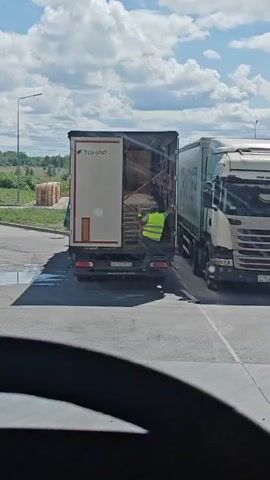 A person wearing a high-visibility vest is seen in the open rear of a white truck. They are reaching into the truck's cargo area. To the right of the white truck is a second truck, a white semi-trailer truck. A forklift, with "TOYOTA" visible on its side, is approaching from the foreground, carrying a pallet of goods. The forklift operator is also wearing a high-visibility vest. The scene appears to be an outdoor loading or unloading area, with a paved surface. In the background, stacks of wooden pallets are visible, along with trees and a cloudy sky. The forklift is moving towards the trucks.