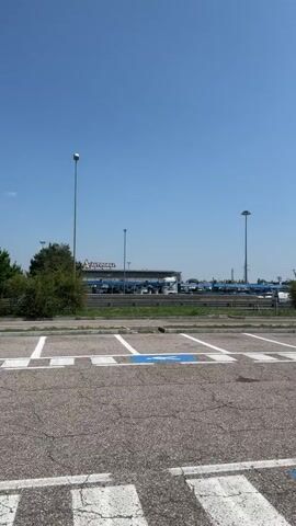 This is a vertical, exterior, daytime video of a parking lot. The camera is positioned low to the ground, facing forward and slightly to the right. The foreground shows a cracked asphalt surface with white and blue painted lines indicating parking spaces. Specifically, there are two handicapped parking symbols visible, each a blue square with a white wheelchair icon. Beyond the parking lot, a road runs horizontally across the frame, with a median strip of vegetation. Several large trucks are parked or driving on the other side of the road. One truck, on the left, is white with black lettering that includes "LECHNER.CZ". Other trucks are blue. Tall lampposts are visible throughout the scene. In the background, a building with a sign that appears to say "AUTOSCUOLA" is present. Traffic signs are visible on the right side of the road, including a yield sign and a "no entry" sign. The sky is clear and blue.