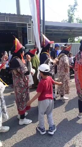 The video shows a group of people, mostly women, dressed in costumes that include conical red and yellow hats and long patterned robes. Some individuals are wearing hijabs. They are standing outdoors on a paved surface, possibly a street, with buildings in the background. A red and white flag is visible in the upper right corner. Several people are holding small red flags or what appear to be candy wrappers. One person in the foreground is looking at their phone. A young boy in a red shirt and gray pants is walking past the group on the right side of the frame. The atmosphere seems to be festive or celebratory.