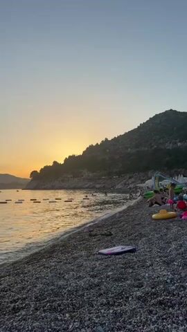 This is a vertical video showcasing a beach scene at sunset in Kamenovo, Montenegro. The sun is setting behind a lush, green hill on the right side of the frame, casting a warm, golden glow across the sky and the water. The sky transitions from a clear, pale blue at the top to a soft orange and yellow gradient near the horizon.

The foreground is dominated by a pebbly beach, with several paddleboards and inflatable water toys scattered on the stones. The water is calm and reflects the sunset's colors. In the sea, there are a few buoys marking a swimming area, and several people are visible enjoying the water, some swimming and others floating. In the distance, more hills form a picturesque backdrop to the bay.

The overall mood is peaceful and serene, with the soft light and gentle waves creating a relaxing atmosphere. The time of day is clearly evening, as indicated by the sunset. There are no visible text overlays in the video.