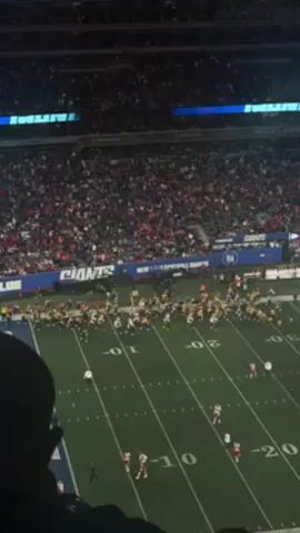 The video shows an American football game from a high vantage point. The field is lined with yard markers and populated by players in yellow and white jerseys and red and white jerseys. Large crowds fill the stadium seats, predominantly wearing red. The sky is dark, suggesting an evening game. The camera is handheld and slightly shaky. A person's head is visible in the bottom left corner, partially obstructing the view. Stadium signage is visible along the sides, including the word "GIANTS."