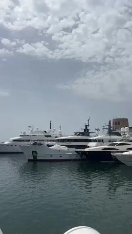 The video shows a panoramic view of a marina filled with large yachts docked in calm, green water. The sky is partly cloudy, with white cumulus clouds against a pale blue background. In the background, a row of white buildings with traditional architecture line the waterfront, featuring balconies and windows. Some buildings have awnings and businesses at street level. Palm trees are visible, adding to the Mediterranean ambiance. A distinctive round tower with a crenellated top is visible on the left side of the frame. The yachts vary in size and design, with several large, luxurious vessels prominently displayed. The camera pans slowly across the scene, showcasing the expanse of the marina and the upscale waterfront. No people are clearly visible on the yachts or along the waterfront.