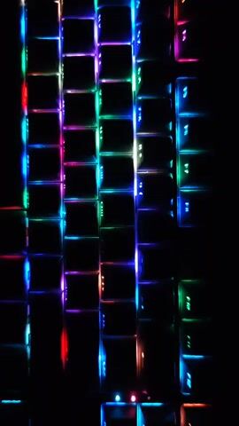The media file is a close-up, top-down shot of a keyboard with multi-colored LED backlighting. The keys are primarily black with illuminated letters and symbols. The lighting shifts through various colors, including blue, green, pink, and red, creating a vibrant and dynamic effect against the dark background. The image is vertically oriented, focusing on the arrangement of the keys. There are no people or discernible environmental details that suggest a specific location, time of day, or weather. The scene is purely focused on the visual aesthetic of the illuminated keyboard. No text is clearly visible on the keys, other than what would typically be found on a keyboard. The overall mood is modern and technological, with the colorful lights creating a somewhat futuristic or gaming-oriented atmosphere. The media file is a still image, with no action or event taking place beyond the visual display of the lights.
