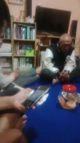 A group of people are sitting on a blue floor. There are several jars of snacks and plates of food on the floor. In the foreground, a person is holding a smartphone, recording the scene. In the background, there is a bookshelf filled with books and some other items. A fan is visible on the wall. The lighting is dim, suggesting an indoor evening setting.