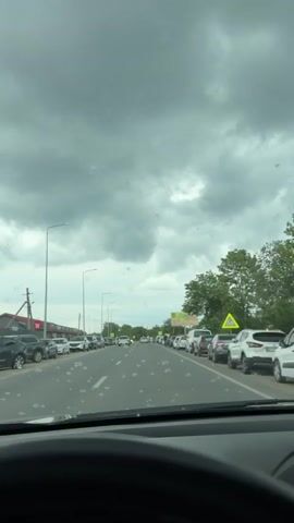 The view is from inside a car, likely the driver's seat, facing forward onto a street with cars parked tightly along both sides and moving slowly in the center lane. The sky is overcast and gray. A man is standing on the right side of the road. The street has street lights, a pedestrian crossing sign, and green trees along its side. The road appears wet or speckled, possibly from a light rain.