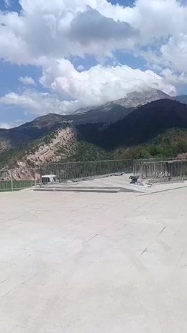 The video shows a mountain landscape with a large flat paved area in the foreground. There are several people working on the paved area, some in orange vests. There are also metal railings and some construction materials. The sky is blue with white clouds.