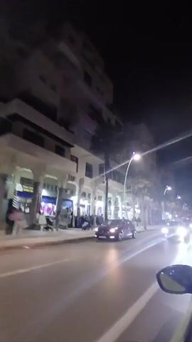 The video depicts a night scene of a city street. The camera, likely mounted in a moving vehicle, captures the view from the driver's side, as cars pass by on the well-lit street. Buildings line the street, with businesses visible on the ground floor. Palm trees add to the urban landscape. Pedestrians can be seen walking along the sidewalks.