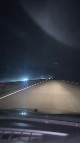 The video shows a first-person view from inside a vehicle driving at night on a highway. The headlights of the vehicle illuminate the road ahead, revealing lane markings and other vehicles in the distance. The sky is dark and appears to be foggy or hazy, obscuring the visibility of the surroundings. The interior of the car, including the dashboard, is partially visible in the foreground. The scene depicts a common nighttime driving scenario on a multi-lane road.