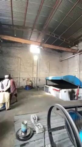 The video shows a large, industrial workspace. The space is dimly lit, with a tall, metal roof structure. There is a large overhead crane system running along a beam. Two men in traditional Pakistani clothing stand near the wall observing the crane. In the foreground is a spool of wire on what appears to be a machine. There's a large blue metal structure in the background, possibly a piece of equipment. The walls are made of light-colored bricks. The general impression is one of a manufacturing or storage facility.