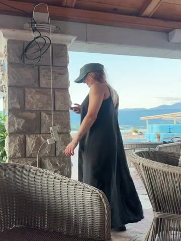 A woman wearing a dark, sleeveless, low-back dress and a green baseball cap stands on a terrace looking at her phone. She has tattoos on her left arm and back. The terrace is made of stone and has wicker chairs. There is a pink flamingo statue on the terrace near the water. In the background, there is a lake and mountains. The sky is clear and blue.