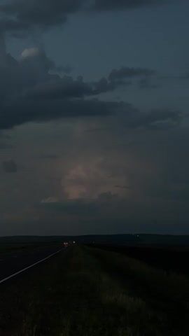 The media file depicts a dark, moody sky over a rural landscape at dusk or nighttime. A road stretches into the distance, lined with dark vegetation. Two cars with their headlights on are visible on the road, suggesting travel. The sky is dominated by heavy, dark clouds, with some lighter, illuminated clouds in the distance, possibly indicating a storm or sunset. The overall mood is somber and atmospheric. The location is identified as Kurgany, Russia. There is no visible text in the media.