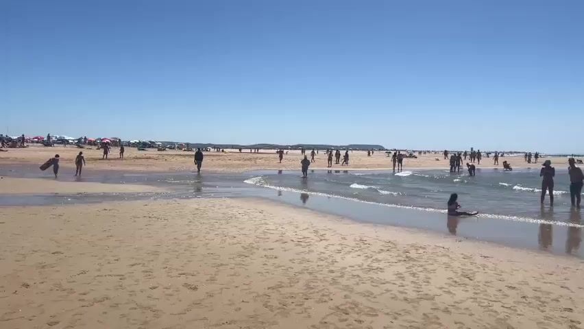 A wide shot captures a crowded beach on a sunny day under a clear blue sky. The foreground is dominated by a sandy beach with gentle waves washing ashore. In the mid-ground and background, numerous people are visible, many wading in the shallow water, swimming, or walking along the shoreline. The sea extends to the horizon, with a few indistinct shapes that might be distant boats. The overall atmosphere suggests a leisurely and popular day at the beach.