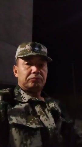 A man in a camouflage uniform and matching hat is looking at the camera. The background is dark with some muted lighting. The man has short, dark hair and a stern expression. The video appears to be a selfie taken in low light conditions.