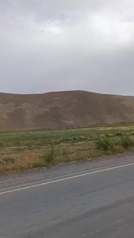 A video shows a road beside a field and a hill in the background. The sky is overcast. A car appears to be moving forward as the road is in the foreground.