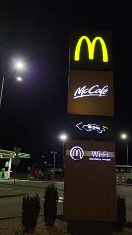 The video opens at night with a view of a McDonald's sign and a BP gas station. A man walks across a pedestrian crossing. The camera then pans to the McDonald's sign which advertises McCafe and Wi-Fi. The video continues to show the gas station and a parked bus in the background. The scene is well-lit by streetlights. The video ends with a view of a parking lot.