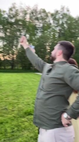 The video shows a group of people gathered on a grassy lawn, possibly at an event or celebration. One man is seen gesturing with his arm raised in the air. Other people in the group are walking and conversing. A person in a yellow shirt is holding a smartphone and appears to be recording or taking photos. The background includes trees, a house, and a light pole. The sky is visible and appears to be dusk or early evening.