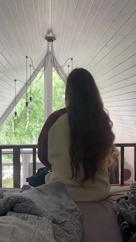 A person with long, dark, wavy hair sits on a bed, facing away from the camera. They are wearing a maroon and cream colored sweatshirt. The person is running their fingers through their hair. The environment appears to be a room with a sloped ceiling. A modern chandelier with multiple bulbs hangs from the ceiling. Through a large window behind the person, trees and greenery are visible. The bed is covered with blankets and pillows.