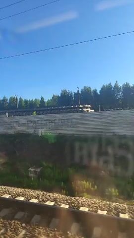 The video shows a view from a moving train. The foreground features train tracks with ballast. Beyond the tracks, there is a grassy embankment. Further in the background, there is a long, grey, slatted structure, possibly a fence or sound barrier. A line of trees forms a backdrop behind this structure, with a clear blue sky above. Power lines are visible against the sky. The lighting suggests it is daytime, with sunlight casting shadows and reflections on the window. There are no people visible in the frame.