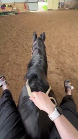 The video is filmed from a first-person perspective, looking forward from the back of a dark-colored horse, likely black or dark bay. The horse is moving at a slow, steady pace, possibly a walk or slow trot, across a sandy or dirt floor within an indoor riding arena. Its mane is neatly braided along its neck.

The rider's lower legs, dressed in black pants, are visible on either side of the horse's barrel. Their feet are clad in light-colored sandals with colorful straps. The rider's left hand, wearing a white smartwatch, holds a light-colored rope, which appears to be part of a halter or bitless bridle used to guide the horse.

The setting is an indoor equestrian facility, characterized by a high ceiling with visible metal support beams. Natural light streams in from a large, open doorway at the far end of the arena, revealing a glimpse of green foliage and a red vehicle in the background, suggesting an outdoor area beyond. The overall scene depicts a calm horseback riding activity in Slashchevo, Russia. No visible text is present.