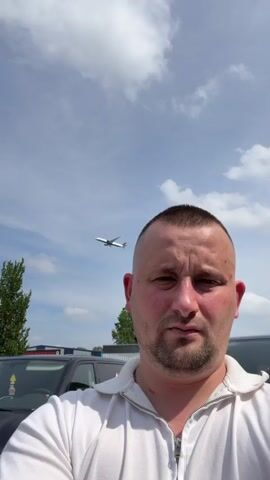A man with a buzz cut and stubble, wearing a white zippered shirt, faces the camera in a selfie-style video. Behind him, a white airplane is visible in the sky. The background also shows trees, cars, a fence, and a street light against a blue sky with white clouds. The man's expression is neutral. The video appears to have been filmed outdoors during the daytime.