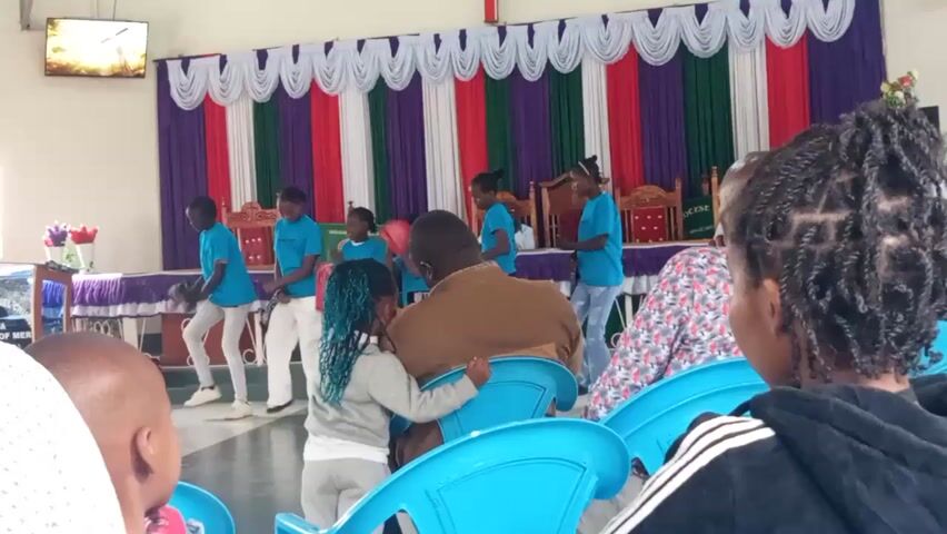 A group of children in matching blue t-shirts and white pants stand on a stage behind a table draped with purple and white fabric. They appear to be performing a dance or play, as some are holding red cloths. The stage is decorated with a backdrop of colorful vertical fabric stripes in white, purple, green, and red. In the foreground, an adult man in a brown jacket is seated, with two young children, one with blue hair, sitting on his lap. Other audience members are visible in the foreground, seated in blue chairs. A large screen is mounted on the wall behind the performers. The environment appears to be an indoor venue, possibly a church hall or community center.
