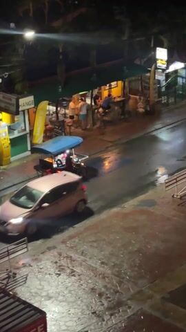 The video shows a street scene at night in what appears to be a tropical location. The street is wet, reflecting the streetlights. A motorized cart with a blue canopy drives away from the camera. The cart appears to be a mobile food or drink vendor. On the side of the street, there is a bar or restaurant with people seated at the counter. There are also advertisements visible. The overall lighting is dim.