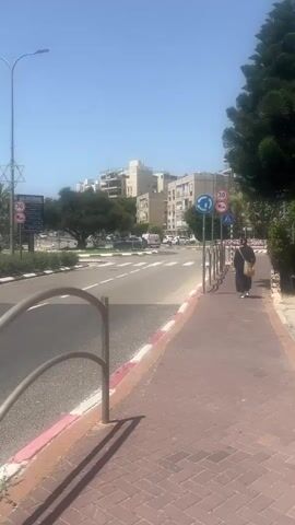 The video shows a street scene on a sunny day. The camera is positioned behind a metal railing on a sidewalk paved with red and white bricks. The street has a pedestrian crossing and a roundabout in the center with greenery. Several vehicles, including white ambulances with red markings, are visible circulating around the roundabout. A street sign with Hebrew lettering and a Star of David symbol is posted nearby. Buildings of various heights and designs line the background.
