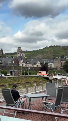 A man sits at a table on a deck overlooking a town with buildings and a church on a hill covered in vineyards. An ambulance is visible driving on a road in the background. The sky is cloudy.